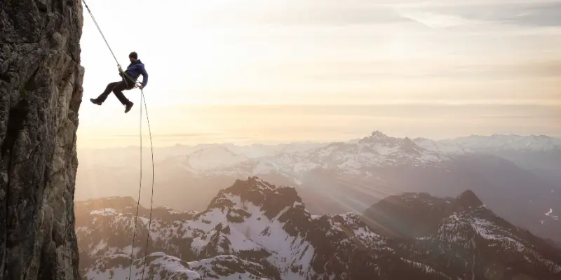 Mental Toughness Rappelling Mountain Cliff Sunset Person rappelling down a mountain cliff at sunset, symbolizing mental toughness and resilience in the face of challenges, with a vast mountain landscape in the background.